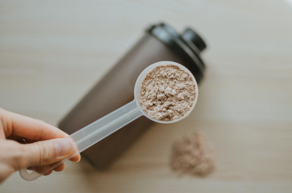Imagem de uma mão a segurar uma colher-medida com pó bege, com um shaker desfocado ao fundo sobre uma superfície clara.