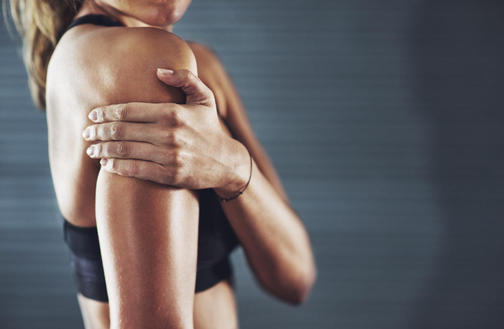 Pessoa em roupa desportiva a segurar o ombro com a mão, sugerindo dor ou desconforto muscular.
