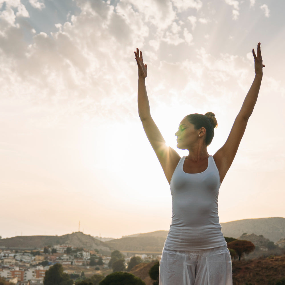 Imagem de uma mulher ao ar livre com os braços erguidos em direção ao céu ao nascer ou pôr do sol, associada a bem-estar, equilíbrio e saúde natural.