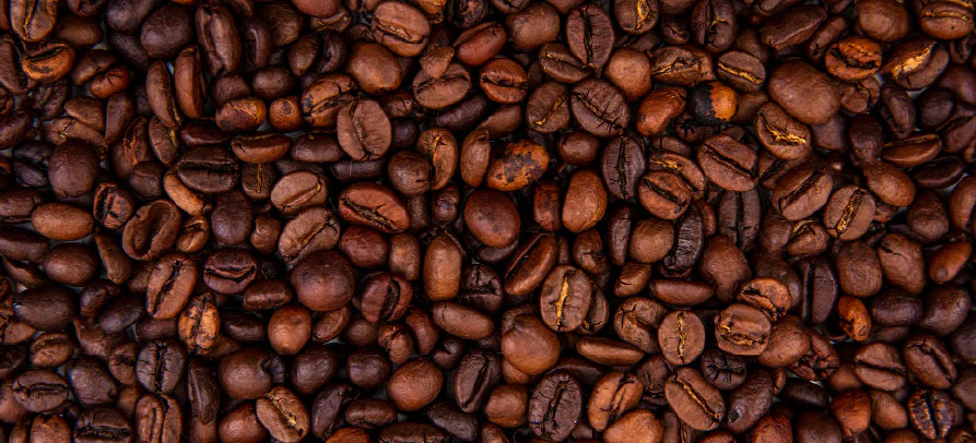 Grãos de café torrados vistos de perto, cobrindo toda a imagem com diferentes tons de castanho e textura detalhada.