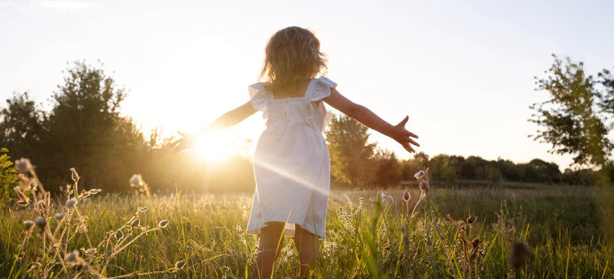 Criança de vestido branco num campo ao pôr do sol, de braços abertos, rodeada pela natureza.