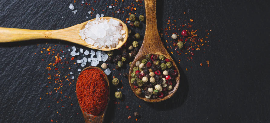 Colheres de madeira com sal grosso, pimenta em grão e especiarias moídas, dispostas sobre uma superfície escura.