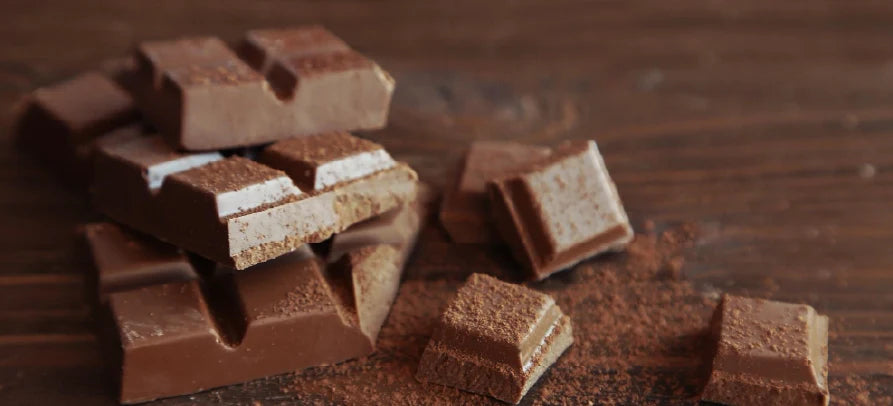 Pedaços de chocolate de leite empilhados e espalhados sobre uma superfície de madeira, com pó de cacau à volta.