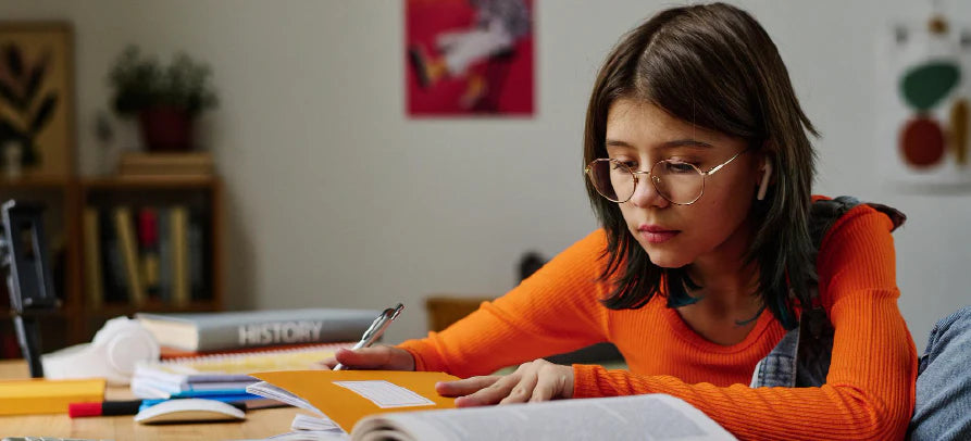 Rapariga de óculos a estudar numa secretária, a escrever num caderno com livros e material escolar à volta.