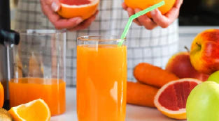 Copo de sumo de laranja fresco com palhinha, servido numa cozinha, rodeado por frutas e legumes como laranjas, toranja, maçã verde e cenouras.