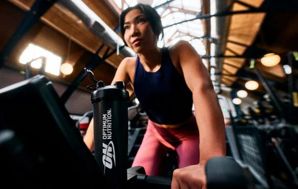 Mulher a treinar numa bicicleta estática no ginásio, inclinada para a frente, com roupa desportiva azul e rosa. Em primeiro plano vê-se um shaker preto da Optimum Nutrition.