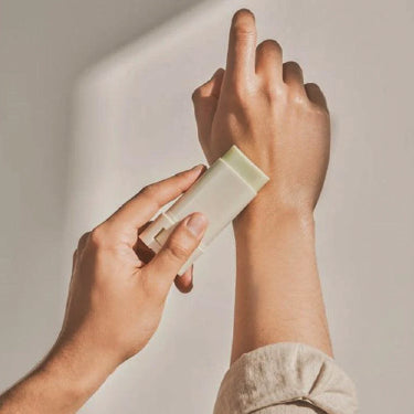 Pessoa a aplicar um produto de skincare em stick no antebraço, num cenário minimalista com luz natural.